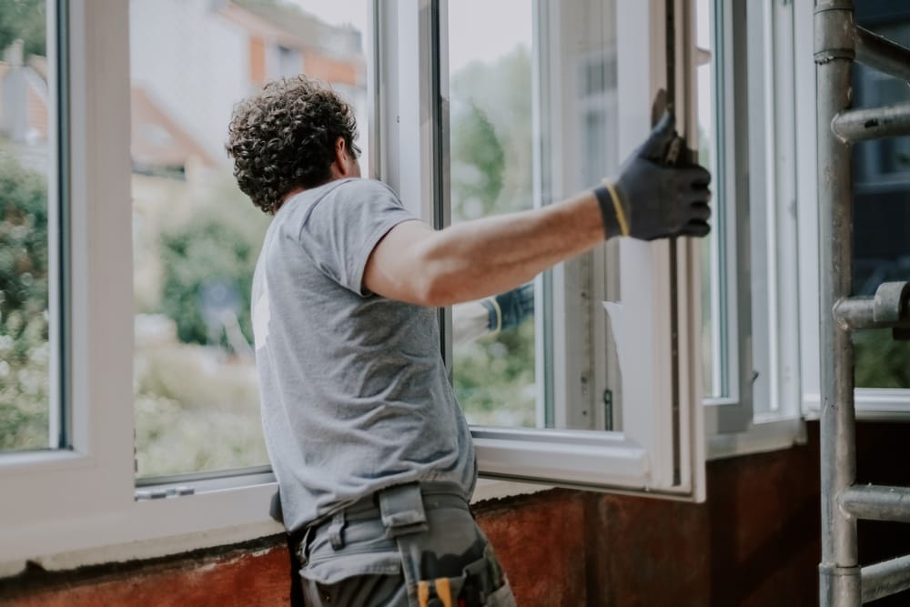 Un jeune homme caucasien, beau, portant un uniforme gris, est vu de dos en train de réparer ou d’installer un cadre de fenêtre dans une maison, vue rapprochée de côté. Concept de rénovation domiciliaire et d’installation de fenêtres.